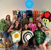 Group photo of people holding various colorful tufted rugs shaped as a tropical leaf, fried egg on toast, rainbow, mushroom, and strawberry design, with yarn cones on the wall behind them and Chillax Studio sign above
