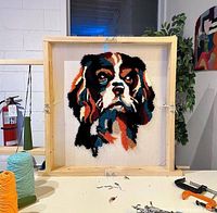 Finished tufted dog portrait displayed in a wooden frame with yarn cones and tufting tools nearby.