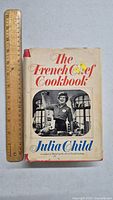 Front cover of The French Chef Cookbook by Julia Child showing book title, author's name, and photo of Julia Child in kitchen setting. A ruler beside the book for scale.