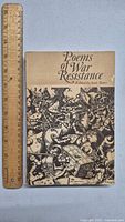 Front cover of the Poems of War Resistance book displayed next to a 12-inch ruler to show size, featuring detailed black-and-white war scene artwork.