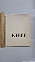 White hardcover book titled 'KIEFF' placed beside a wooden ruler for size reference.