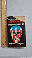 Front cover of Fear and Loathing on the Campaign Trail '72 paperback book showing title, author, and skull illustration with U.S. flag helmet. Includes ruler for size reference.