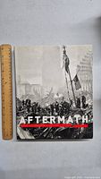 Front cover of 'Aftermath' book showing black and white photo of 9/11 aftermath with city ruins and American flag, with ruler for size reference.