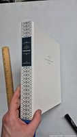 Hand holding white hardcover book showing front cover design and general size with ruler for scale.