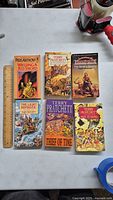 Top-down photo of six fantasy paperback books arranged on a flat surface with a ruler for scale. Various titles by Terry Pratchett and others shown.