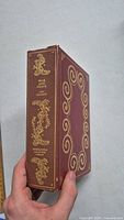 Side-angle view of War and Peace book showing spine and part of front cover with gold embossed decorations and lettering on reddish-brown cover