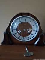 Front view of the antique wooden mantel clock with visible clock face, Roman numerals, and winding key placed in front.