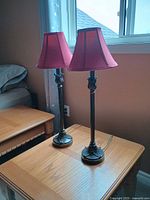 Two bedside lamps standing on a wooden surface near a window, showing overall size, shape, and color of lamps and shades.