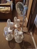Five oscillating fans grouped with four on the floor and one standing fan next to a window. White color, all have front safety grills and power cords visible.