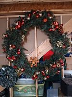 Photo of a large outdoor Christmas wreath segmented into 4 pieces, decorated with red and gold Christmas ornaments and ribbons, resting on a wooden frame in an indoor storage area.