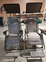 Pair of brown mesh outdoor lounge chairs with adjustable top shade canopies, visible headrests, footrests, metal frames, and armrests standing side by side indoors.