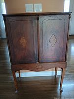 Front view of antique wooden secretary desk showing closed dual cabinet doors with decorative inlay and the legs with carved apron below.