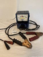 Front angled view of the blue battery tester showing meter face, cables with clips and probes on white background.