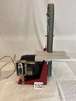 Side profile of bench belt sander showing red metal frame, aluminum table, black motor, and sanding belt.