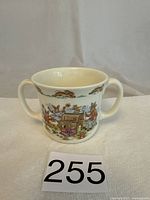 Front view of the two-handle white porcelain Bunnykins mug with colorful bunny character illustrations surrounding it, featuring rabbits near a mailbox and in outdoor scenes.