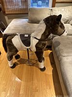 Side view of plush toy horse showing brown and white coloring with saddle, bridle, and black hooves standing on floor next to sofa.