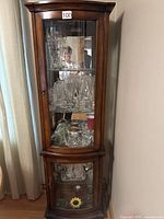 Front view of a tall, wooden corner curio cabinet with two glass doors and visible glass shelves, shown with glassware inside.