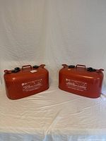 Two red metal gasoline cans with black caps and handles, positioned side by side on a white cloth background.