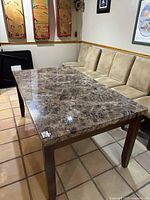 Dining table with faux marble pattern top and wooden legs placed on a tiled floor, surrounded by beige faux suede chairs.
