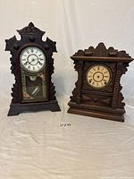 Front view of two vintage wooden table top clocks showing their ornate and carved wooden cases. One clock has a white dial with a pendulum visible behind glass, the other has a beige dial. Both show wear.