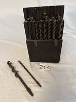 Full view of twelve large vintage brace bits neatly stored in a wooden holder case with two loose bits laid in front to show size and design.