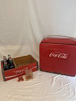 Full display of six Coca-Cola bottles in wooden crate next to red metal Coca-Cola cooler with two bottle caps in front.