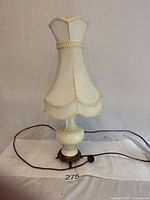 Front angle view of the vintage table lamp showing cream fabric scalloped lampshade, white ceramic base, ornate aged brass base, power cord, and on/off switch.