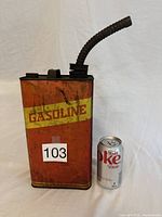 Front view of vintage red metal gasoline can with yellow 'GASOLINE' label, showing flexible black spout and placed next to a soda can for size reference.