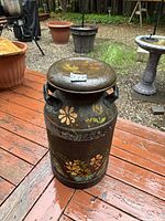Full view of vintage painted metal milk can on outdoor wooden deck, showing floral designs and rustic patina.