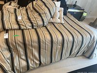 Bed with striped beige, black, and cream bedspread laid over it, along with coordinating pillows showing the overall style and color scheme of the linens and pillows.