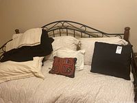 Multiple pillows (black, cream, multicolored) arranged on a bed with cream quilted bedspread and black metal frame.