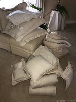 Multiple beige and cream colored pillows and cushions stacked on a sofa and on the floor