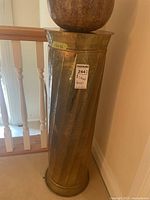 Full view of the tall brass column style stand placed near a staircase, showing cylindrical shape and hammered texture.