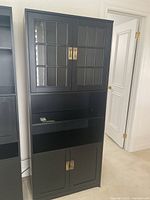 Front view of the tall black cabinet showing two upper glass panel doors, open middle shelf, and two lower solid doors with brass handles.