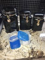 Three black ceramic canisters with metal clasp lids and four plastic food storage containers with blue lids on granite countertop.