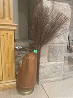 Copper coal scuttle placed against a stone fireplace and wooden furniture. Shows full height and shape with twig bundle inside.