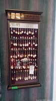 Full view of wood and glass display case with about 40 collector spoons mounted on red fabric backing