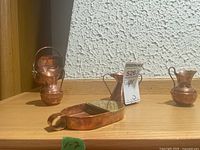 Three small copper miniatures on shelf: two small pitchers and a rectangular tray with handle.