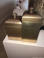 Two square gold-tone jars with lids placed side by side on a white surface, showing the taller (12 inch) and shorter (10 inch) jars with ornate finial tops.