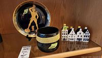 Black and gold Greek art plate, black jeweled bowl, and four Delft KLM ceramic houses on wood shelf