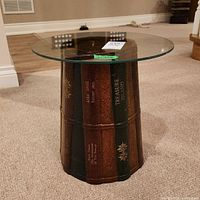 Photo showing round glass top over a cylindrical base designed to look like a stack of antique books with titles and decorative elements in brown and green tones.
