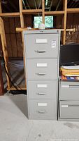 Front view of a tall, light gray, metal filing cabinet with four drawers. Each drawer has a handle and white label holder. Cabinet is standing on a concrete basement floor with a wooden frame wall behind.