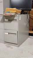 Metal two drawer filing cabinet seen from front and side view, with papers and monitor on top.