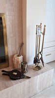 Photo of fireplace hearth showing brass fireplace tools on stand, bellows, pillar candles, and decorative items arranged next to fireplace.
