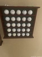 Wooden golf ball display case with 25 golf balls arranged on 5 shelves, showing full lot.