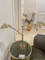 Full view of the brass toned double-arm lamp placed on a round glass table, showing the structure and styling.