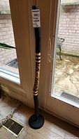 Full length view of the didgeridoo standing on its black round stand near a glass door, showing the entire instrument and mouthpiece with a label attached at the top.