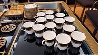 Overview of 14 white cups with black and gold band and a stack of matching saucers on a black glass table