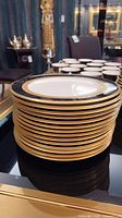 Stack of 14 white porcelain plates with black and gold rims on a glass-topped table.