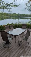 Six outdoor metal frame dining chairs with mesh seats and backs surrounding a glass outdoor dining table on a wood deck overlooking a lake.